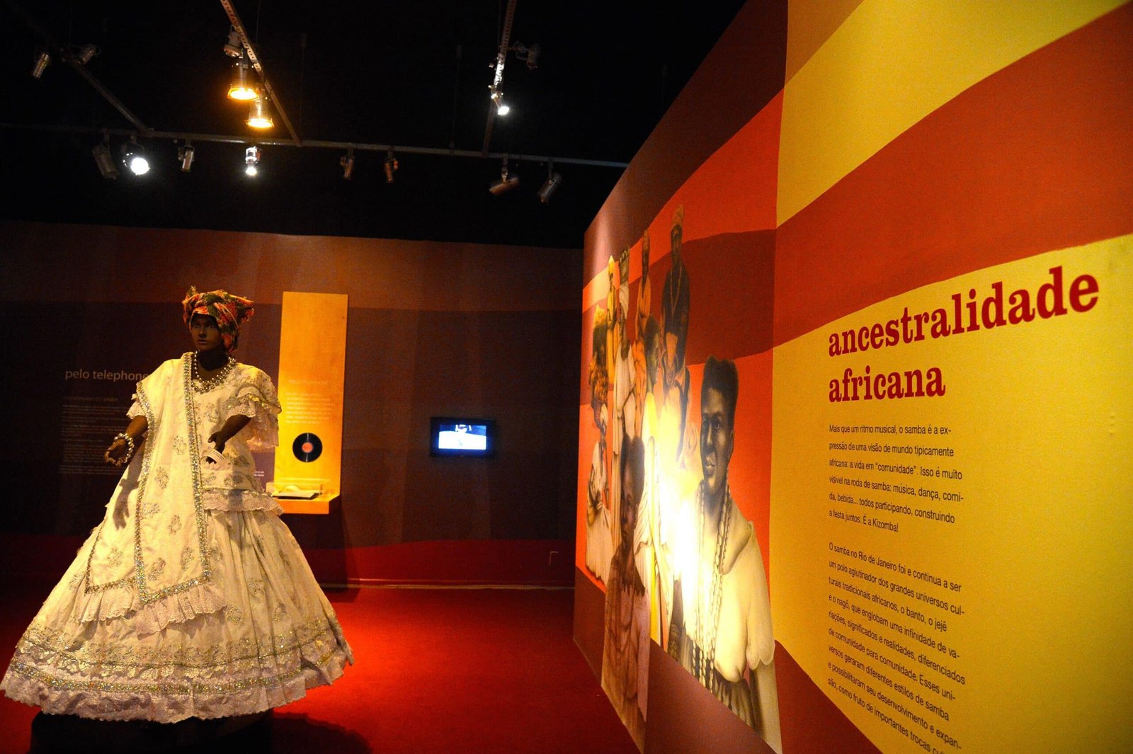 Uma pessoa vestida de branco tradicional está em uma exposição de museu sobre ascendência africana, com painéis informativos vibrantes expostos. Imagem para ilustrar o post sobre museus no Rio de Janeiro.