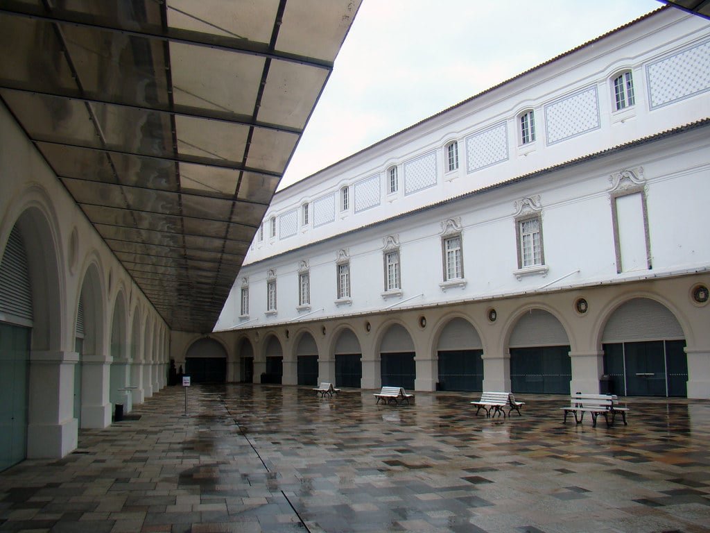 Pátio vazio do Museu Histórico Nacional com prédios brancos em arco e bancos. O chão está molhado, e uma passarela coberta fica à esquerda.