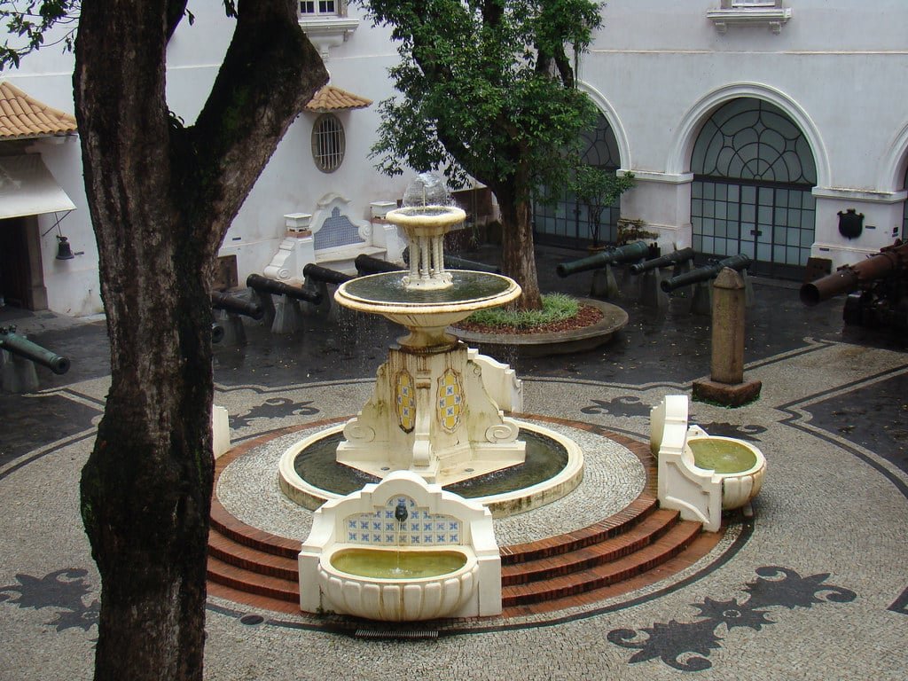 Pátio do Museu Histórico Nacional com uma fonte central, cercado por canhões e árvores. O chão é pavimentado com padrões de pedra intrincados. Imagem para ilustrar o post sobre museus no Rio de Janeiro.