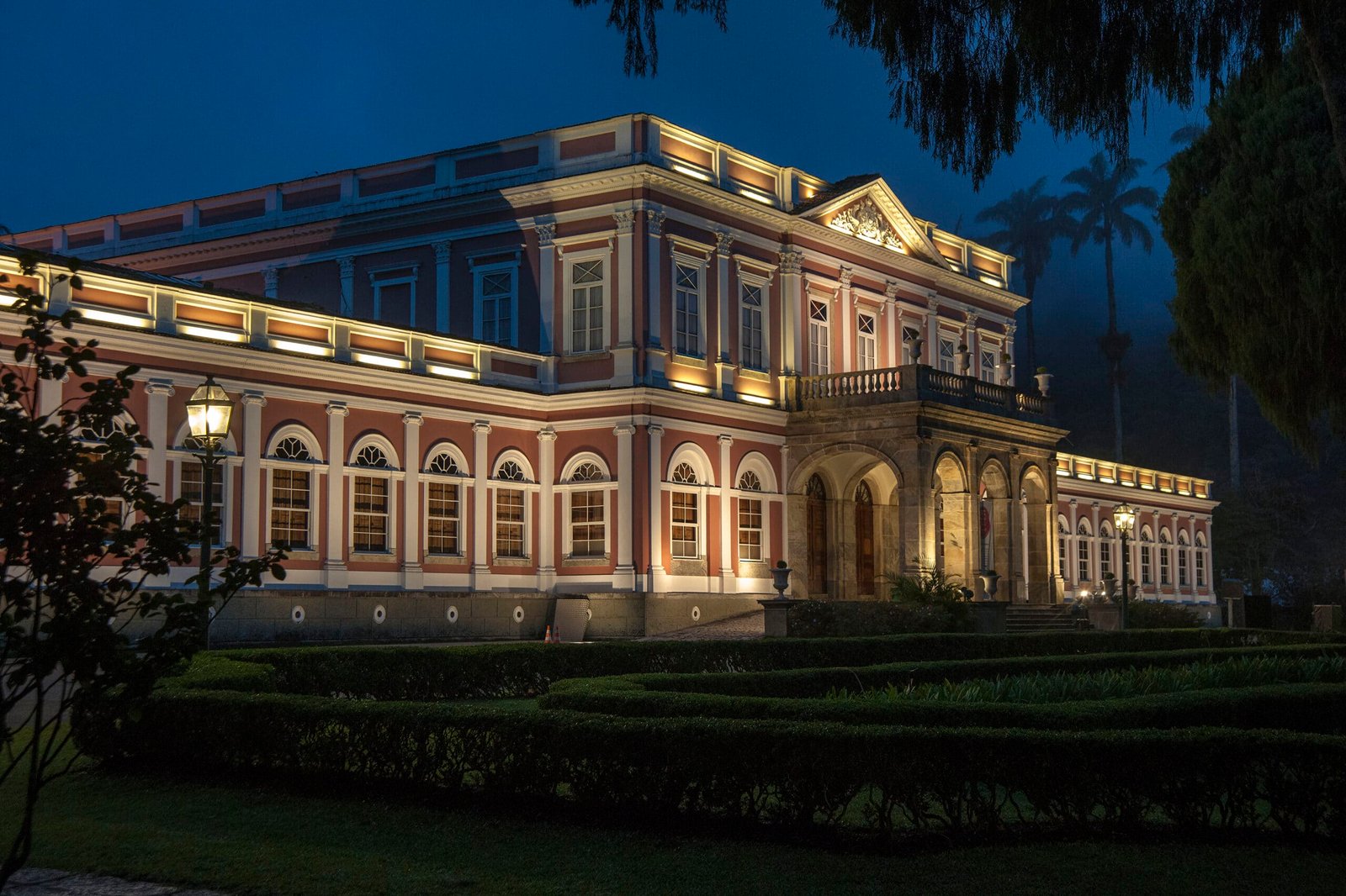 Museu Imperial iluminado com arquitetura clássica fica à noite, cercado por sebes e árvores bem cuidadas. Imagem para ilustrar o post sobre museus no Rio de Janeiro.