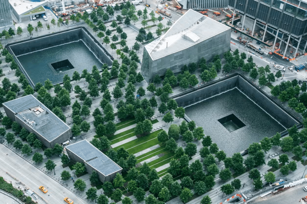 Vista aérea do Memorial do 11 de Setembro na cidade de Nova York, com duas grandes piscinas quadradas cercadas por árvores e edifícios.