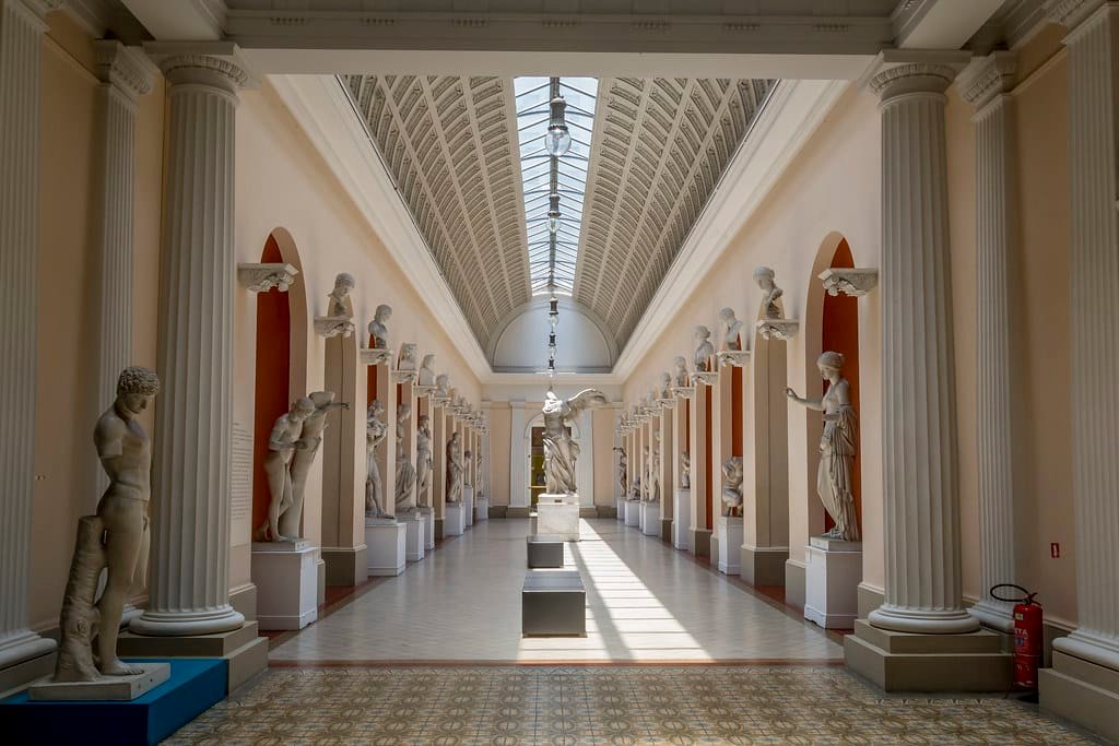 Museu Nacional de Belas Artes com uma galeria iluminada pelo sol com estátuas clássicas alinhadas em ambos os lados, colunas altas e um teto de vidro, criando uma atmosfera iluminada.