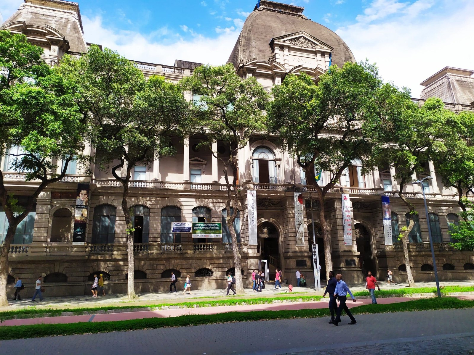 Museu Nacional de Belas Artes com altas janelas em arco e árvores na frente, pessoas caminhando na calçada sob um céu parcialmente nublado. Imagem para ilustrar o post sobre museus no Rio de Janeiro.