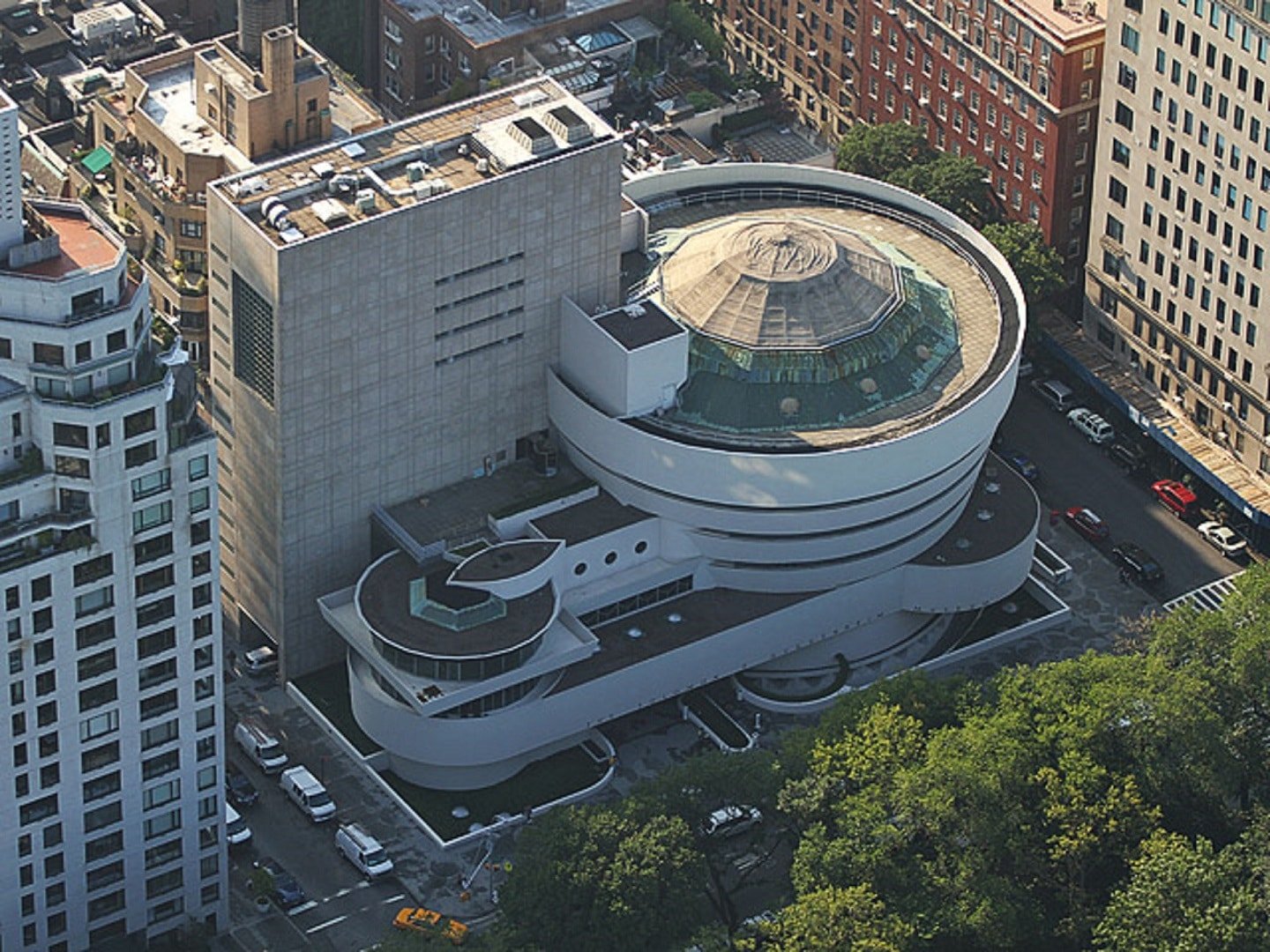 Vista aérea do Museu Solomon R. Guggenheim em formato de espiral com uma seção circular abobadada, cercado por ruas da cidade e vegetação. Imagem para ilustrar post sobre museus em Nova York.