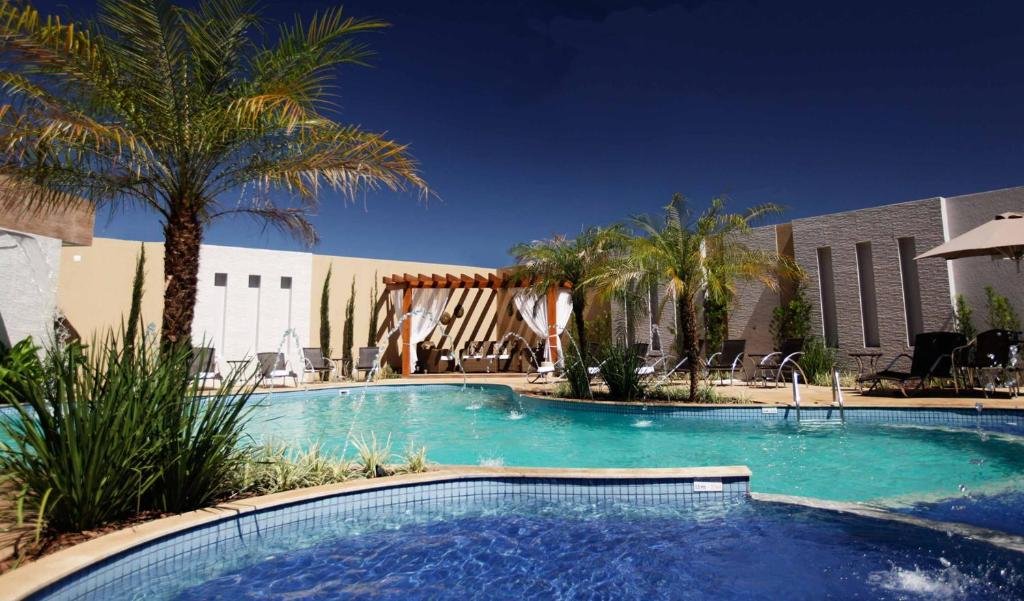 Parte do Nadai Confort Hotel e Spa que mostra a área da piscina ao ar livre com algumas cascatas de água. Em volta da piscina tem algumas plantas e palmeiras verdes, algumas cadeiras e parte da construção do resort atrás. Imagem ilustrando post Resorts em Foz do Iguaçu.