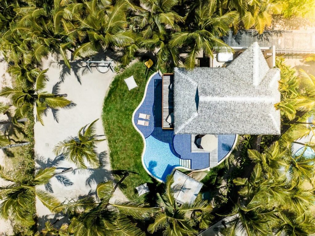 A imagem mostra uma vista aérea de uma área do NANNAI Muro Alto cercada por palmeiras. No centro, há uma piscina de formato orgânico com revestimento azul, adjacente a um bangalô com telhado de palha. À esquerda, uma área de areia com algumas palmeiras e uma rede de balanço, proporcionando um ambiente relaxante. Ao redor, a vegetação é exuberante e verde, destacando o cenário tropical e paradisíaco. Ao lado da piscina, há espreguiçadeiras e um guarda-sol, indicando um local ideal para descanso e lazer.