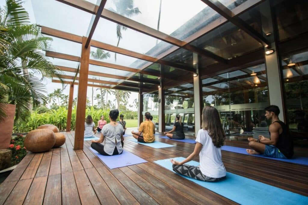 A imagem mostra uma aula de ioga ao ar livre em uma varanda de madeira no NANNAI Muro Alto, coberta por um teto de vidro. Várias pessoas estão sentadas em tapetes de ioga, meditando em posição de lótus, voltadas para o instrutor. A varanda é cercada por vegetação tropical exuberante, com palmeiras e outras plantas ao fundo. Há também grandes esculturas de pedra decorativas ao lado da varanda, criando um ambiente tranquilo e harmonioso para a prática de ioga.