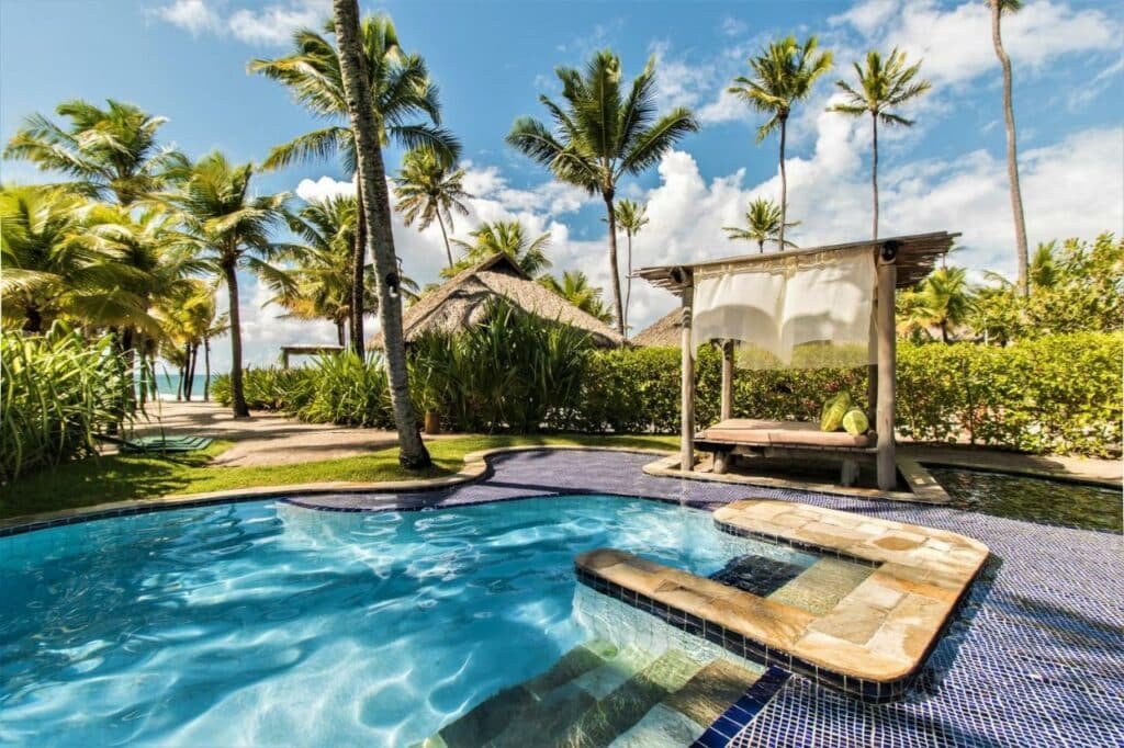 A imagem mostra uma piscina em do NANNAI Muro Alto. A piscina tem bordas arredondadas e é revestida com azulejos azuis. Ao lado da piscina, há um gazebo de madeira com um colchão e travesseiros decorativos, coberto por uma cortina branca. A área ao redor está cercada por vegetação exuberante, incluindo palmeiras e arbustos, criando um ambiente relaxante e privativo. Ao fundo, é possível ver mais palmeiras e um caminho que leva à praia, que está visível ao longe. O céu está azul com algumas nuvens, complementando o cenário paradisíaco.