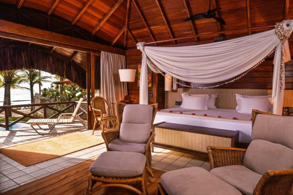 A imagem mostra um quarto do NANNAI Muro Alto, um dos melhores resorts all inclusive no Brasil. O teto é alto e de madeira, com vigas expostas. A cama tem um dossel com um tecido leve e decorativo. Há uma área de estar com poltronas de vime acolchoadas e uma mesa. Ao fundo, há portas de vidro que se abrem para uma varanda com cadeiras de sol e uma piscina privativa. A varanda oferece uma vista para a vegetação tropical e o mar, proporcionando um ambiente relaxante e paradisíaco.