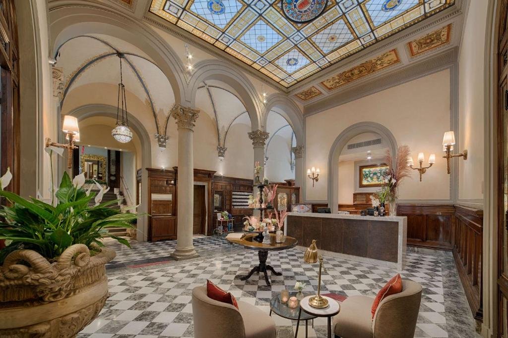 Recepção do NH Collection Firenze Porta Rossa, com decoração clássica, escada dando acesso ao segundo andar, mesa redonda com flores em cima, poltronas e balcão com computador