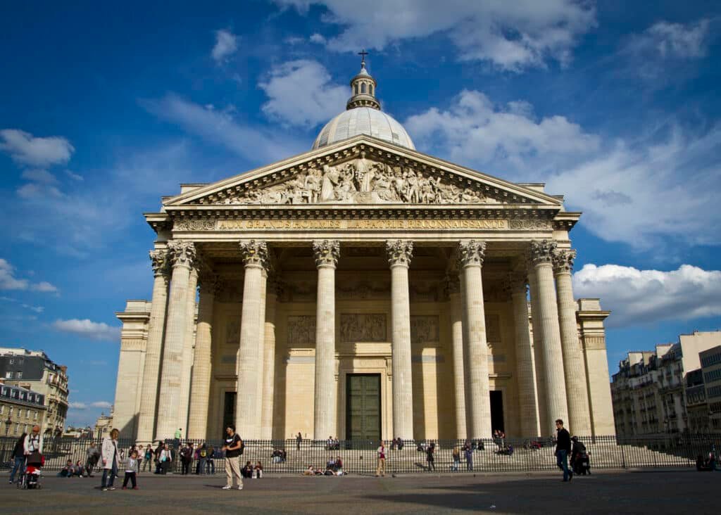 Fachada do Panteão, uma das opções de o que fazer em Paris. Várias colunas e figuras romanas fazem parte do local, e há um domo no topo. Há algumas pessoas passeando ao redor.