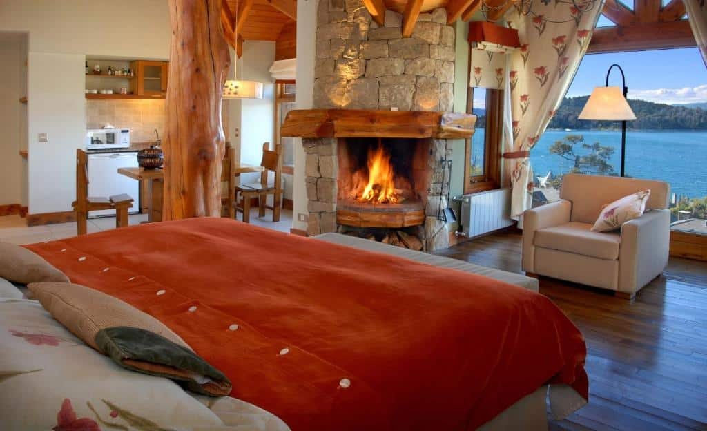 Quarto do Nido del Cóndor Hotel & Spa. Uma cama de casal do lado esquerdo, uma parede de vidro do lado direito, uma poltrona e um abajur e uma lareira.