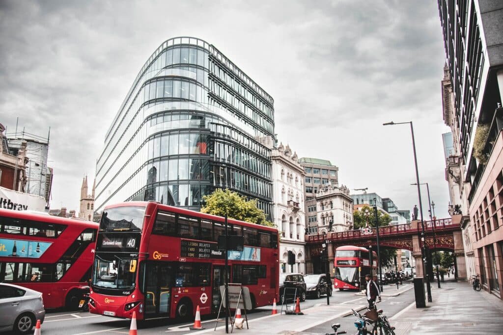 Vista de uma rua de Londres com os famosos ônibus vermelhos, pessoas andando, carros e alguns prédios ao redor