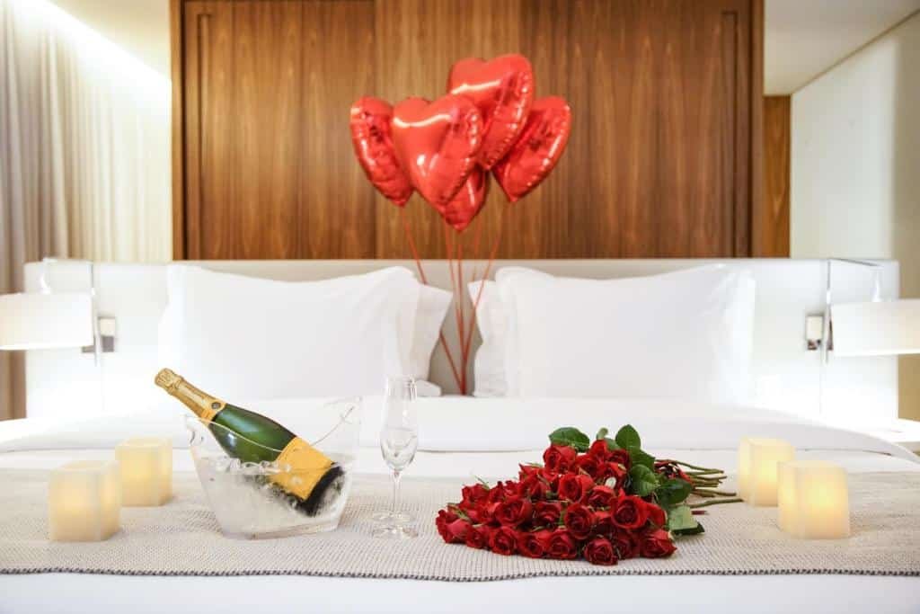 Uma cama de hotel com balões de coração vermelho, um buquê de rosas vermelhas, uma garrafa de champanhe no gelo e velas em um suporte para representar um dos melhores lugares para lua de mel