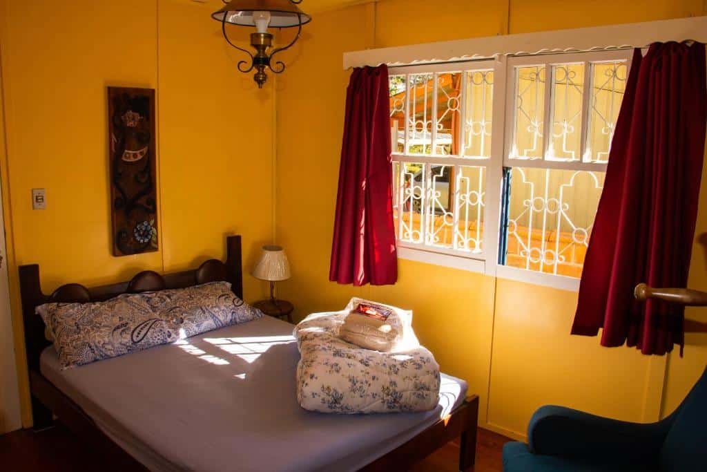 Foto de um quarto no airbnb Nossa casa em Canela. A cama é de casal, e encosta na parede. No seu lado direito, há uma mesa de cabeceira pequena com abajur e uma janela com vista para o quintal. Em cima da cama há roupas de cama. No canto inferior direito da foto há um sofá.