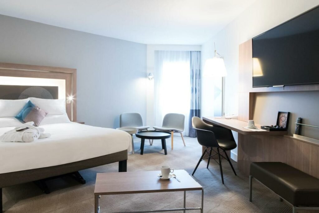 Quarto espaçoso do Novotel Château de Versailles, um dos hotéis perto do Palácio de Versalhes. Uma cama de casal co roupões aos pés está encostada na parede ao lado esquerdo. Logo em frente há uma mesinha com xícara, colher e pires, ao lado da parede onde está um banco, uma TV e a mesa de trabalho com cadeira e abajur. Ao fundo do quarto fica uma janela com cortinas e uma área de estar com poltronas e uma mesinha de centro.