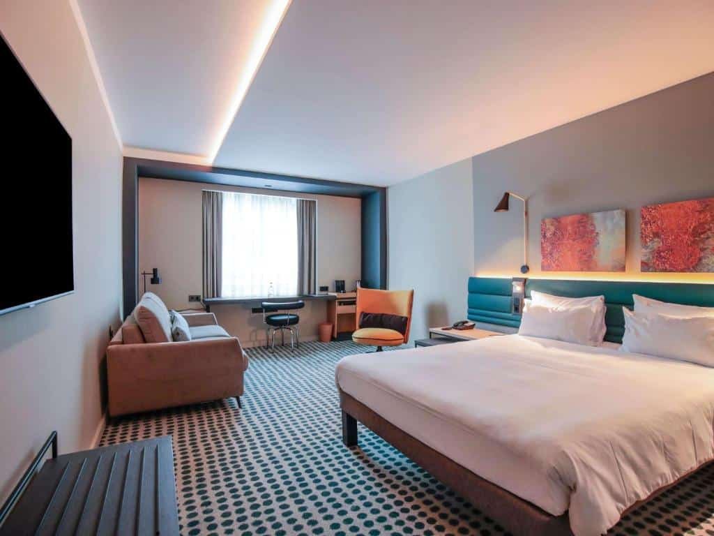 Um quarto espaçoso no Novotel Luxembourg Kirchberg, para ilustrar o post sobre hotéis em Luxemburgo. A cama é extra-grande, box e de casal, e está na direita da foto. Atrás dela, há uma cabeceira azul marinho e dois quadros com artes em tom de coral decorando a parede. Há uma luminária fixada na parede, perto da cabeceira, no lado esquerdo da cama. Na frente da cama, há uma TV de tela plana. Prosseguindo ao fundo do quarto, há um sofá encostado na parede esquerda, a mesma que tem a TV, e, ainda mais ao fundo, há uma janela e abaixo dela há uma escrivaninha preta, uma cadeira e outra pequena mesa de madeira com amenidades em cima.