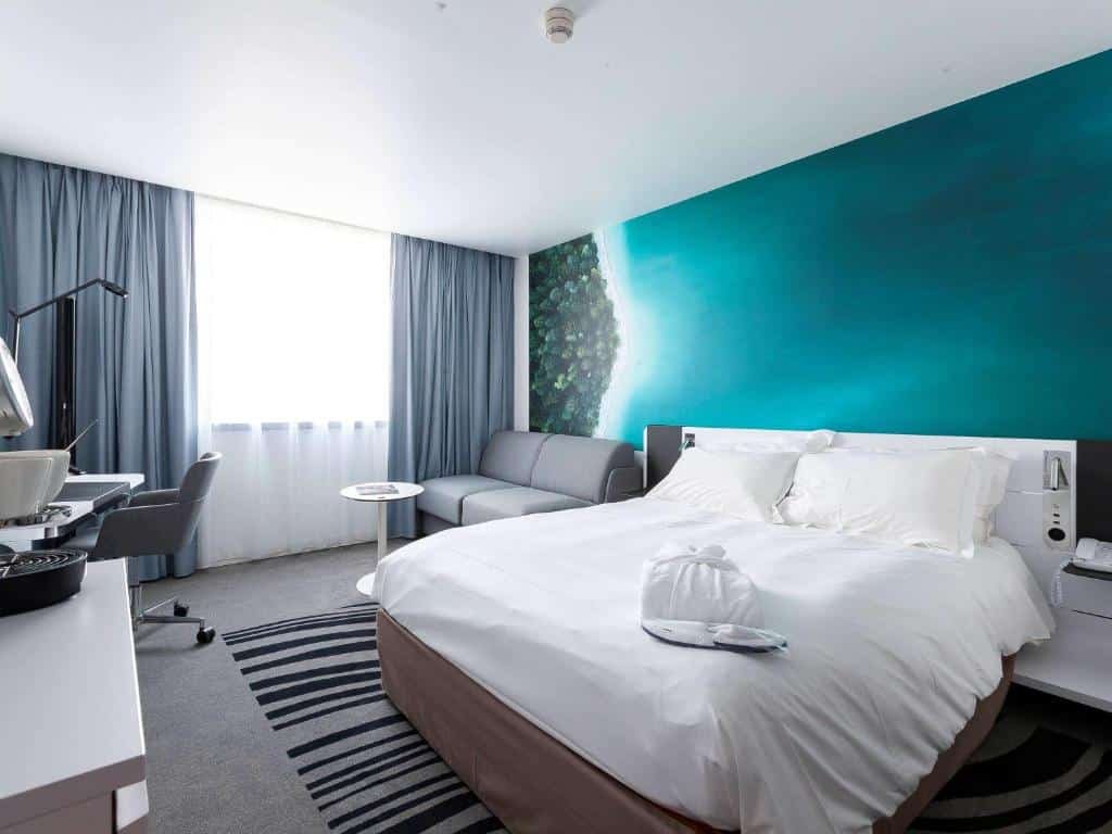 Suíte no Novotel Luxembourg Centre, para ilustrar o post sobre hotéis em Luxemburgo. A cama box de casal está no lado direito da foto, a parede atrás possui uma tintura do mar, em azul marinho. No lado esquerdo da cama, há um sofá pequeno cinza, uma mesa redonda em sua frente e uma janela ao lado. Na frente da cama, vemos de relance um móvel branco com objetos em cima, e no canto do quarto, há uma escrivaninha com uma cadeira giratória, que estão do lado da janela.