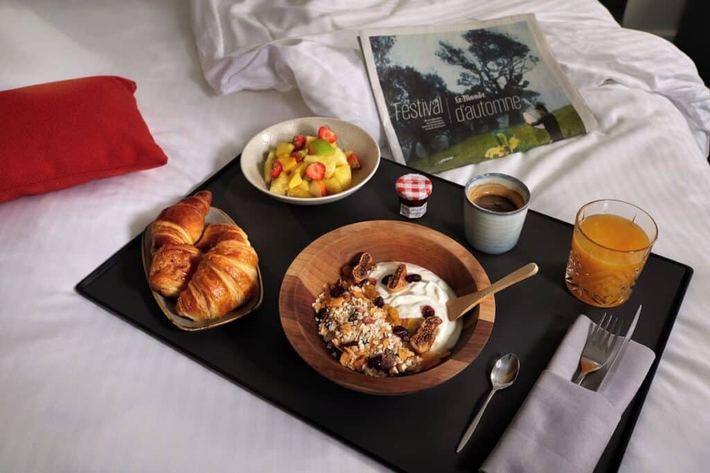 A imagem mostra uma bandeja de café da manhã colocada sobre uma cama com lençóis brancos no Novotel Paris Les Halles. Na bandeja, há um prato com dois croissants, uma tigela de iogurte com granola e frutas secas, uma tigela de frutas frescas com pedaços de morango e abacaxi, uma xícara de café, um copo de suco de laranja e um pequeno pote de geleia. Ao lado da bandeja, há um jornal aberto com a manchete "Festival d