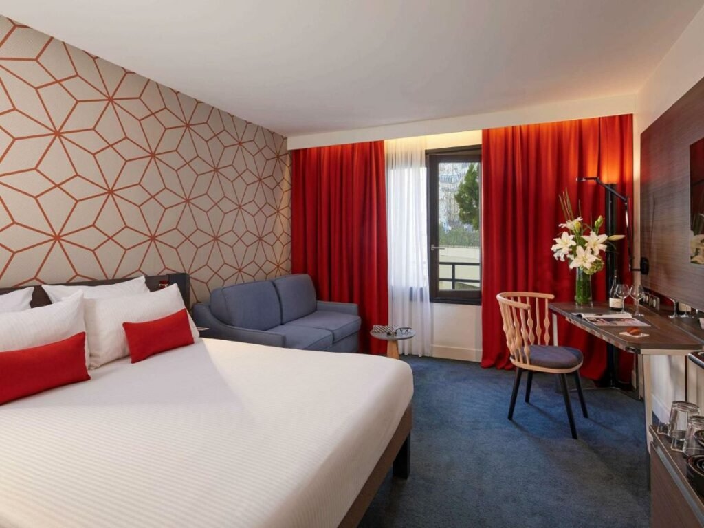 Quarto do Novotel Paris Les Halles, um dos hotéis com café da manhã em Paris. Uma cama de casal está encostada na parede do lado esquerdo e conta com mesinhas de cabeceira e luminárias dos dois lados. Ao fundo do quarto fica uma área de estar com sofá e mesinha de centro na mesma parede da cama, e logo em frente há uma mesa de trabalho com cadeira, luminária, vaso de flores, garrafa de vinho e duas taças de vidro. Em frente à cama há uma TV na parede. Janelas cobertas por cortinas ficam ao lado do sofá.