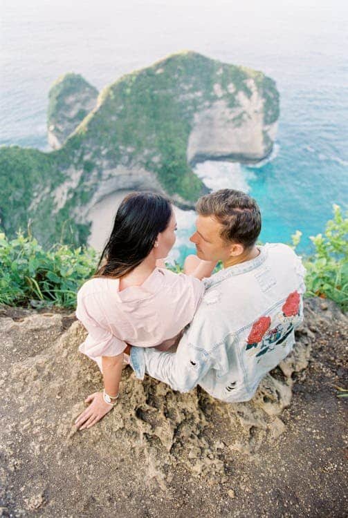 Um casal está sentado na beira de um penhasco com vista para o oceano e o litoral rochoso, com vegetação exuberante ao redor.