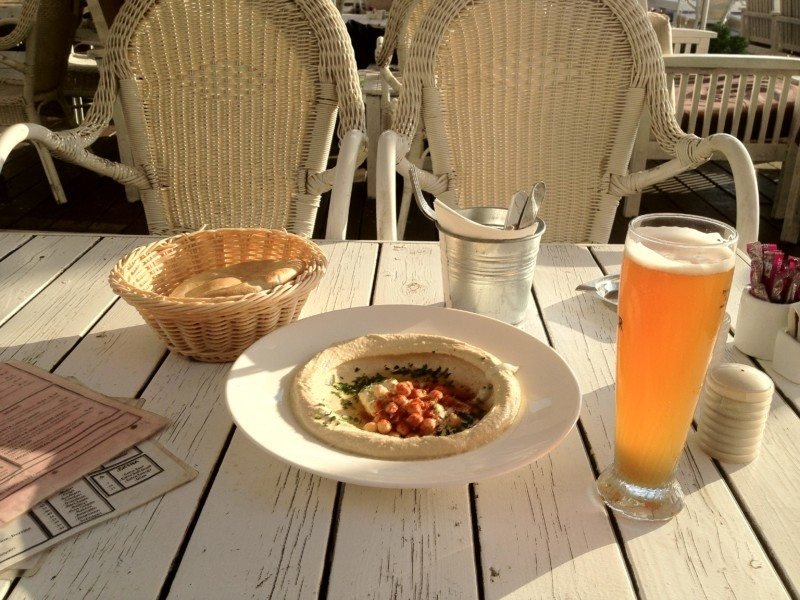 Uma mesa de madeira branca com um prato em cima contendo um alimento israelita, feito de massa e verduras. Há um copo longo de chopp ao lado direito. Há outras coisas na mesa como uma cesta com uma espécie de pão dentro, sal e outros temperos. Ilustra o que fazer em Israel.