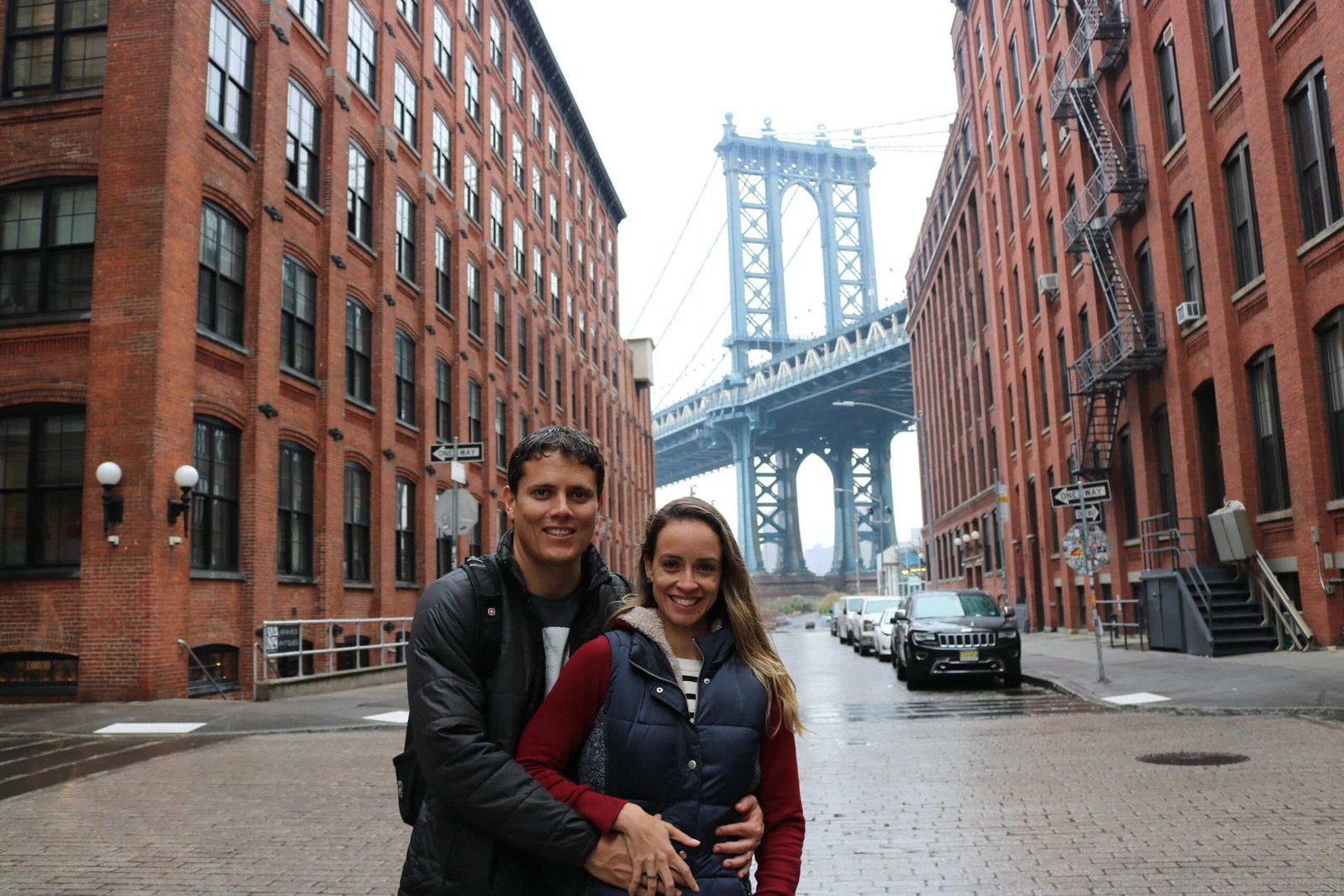 A imagem mostra duas pessoas de pé no meio de uma rua, com prédios em ambos os lados. Ao fundo, é possível ver a Ponte de Manhattan. 