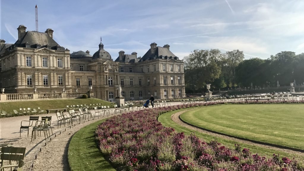 Palácio de Luxemburgo com gramados bem cuidados, canteiros de flores cor-de-rosa e cadeiras verdes espalhadas sob um céu azul claro. Esse é um dos pontos turísticos de Paris.