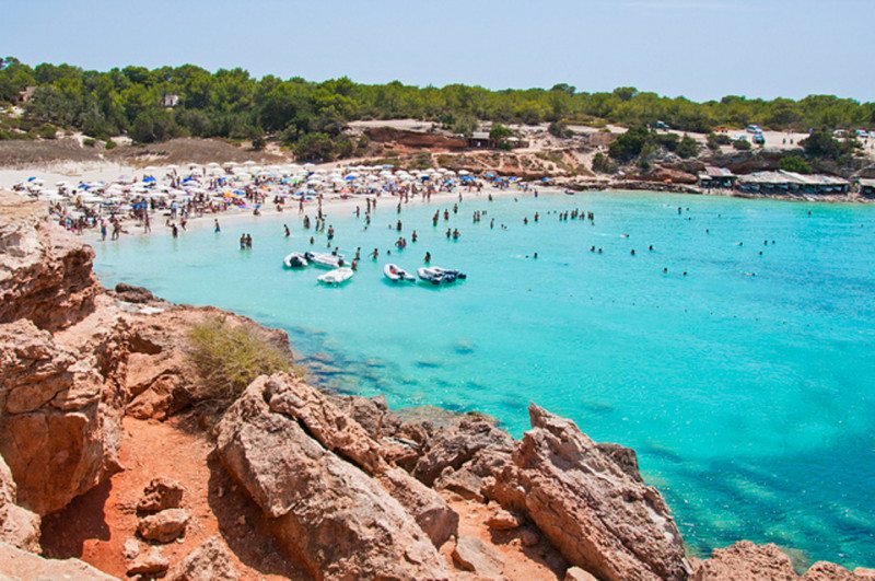 Cala Saona lotada com pessoas nadando em águas azul-turquesa, barcos ancorados e um primeiro plano rochoso com vegetação exuberante ao fundo.