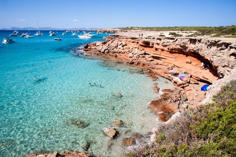 Uma praia rochosa com águas cristalinas azul-turquesa, pessoas nadando e barcos ancorados ao longe, sob um céu azul claro. Essa é a Cala Saona, uma das opções de o que fazer em Formentera.