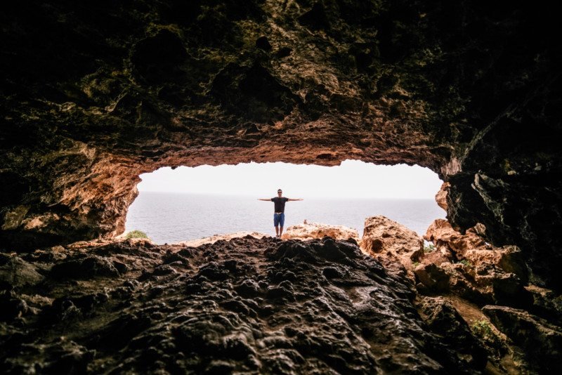 Uma pessoa está de pé com os braços estendidos em uma caverna rochosa, de frente para o oceano visível através da abertura da caverna. Essa é a Cap de Barbaria, uma das opções de o que fazer em Formentera.