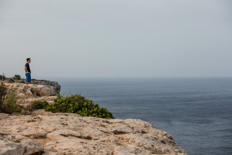 Pessoa em pé na beira de um penhasco rochoso, olhando para o vasto oceano sob um céu nublado. Essa é a vista perto do Farol de la Mola, uma das opções de o que fazer em Formentera.