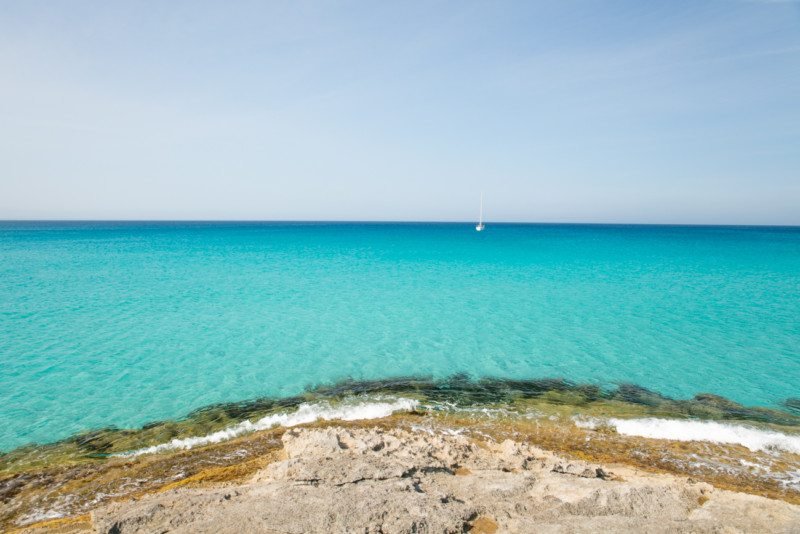 Um mar azul-turquesa claro com um veleiro solitário ao longe, em contraste com um céu azul, visto de uma costa rochosa na Es Calo de Sant Agustin, uma das opções de o que fazer em Formentera.