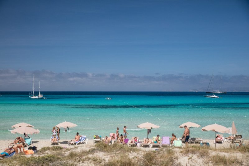 Pessoas relaxam sob guarda-sóis na Praia de Ses Illetes, que é uma das opções de o que fazer em Formentera e tem areia com águas azul-turquesa e veleiros ao longe, sob um céu azul claro.