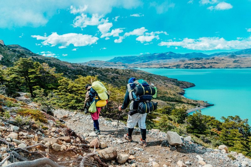 Dois caminhantes com mochilas caminham por uma trilha rochosa em direção a um lago azul-turquesa na Torre del Paine, na Patagônia Chilena, cercado por vegetação e montanhas distantes sob um céu azul para representar um dos melhores lugares para lua de mel