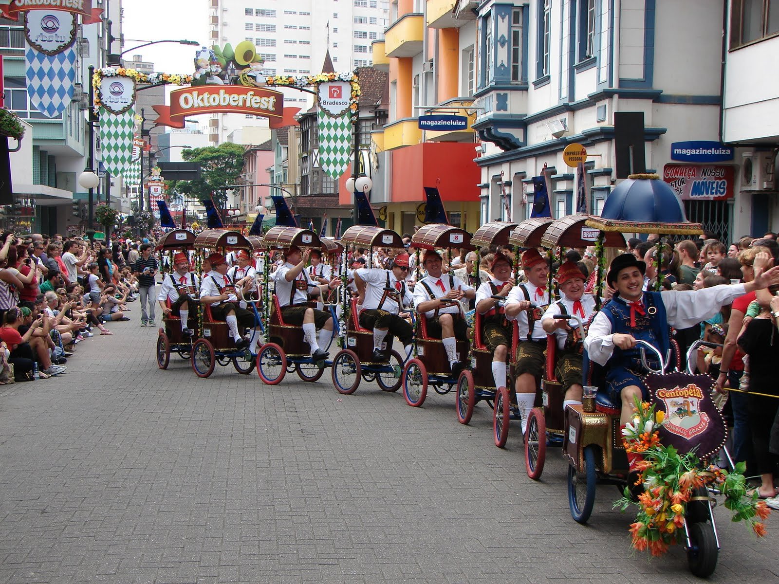Um grupo de pessoas em trajes tradicionais anda de bicicleta com vários assentos por uma rua, com multidões assistindo durante o Oktoberfest Blumenau.