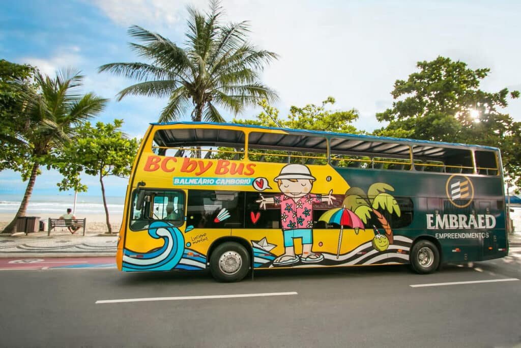 Um ônibus de turismo todo colorido andando, atrás palmeiras e a orla da praia.