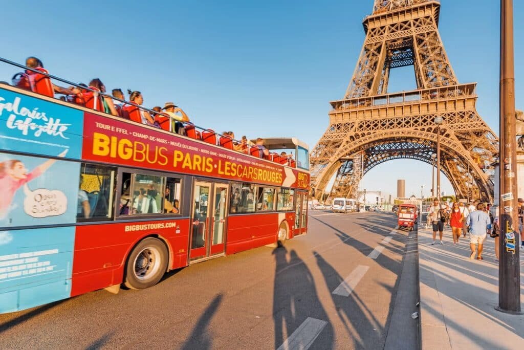 Um ônibus turístico vermelho de dois andares passa perto da Torre Eiffel, em Paris, com passageiros no andar superior e pessoas caminhando nas proximidades para ilustrar o post de passeios em Paris.