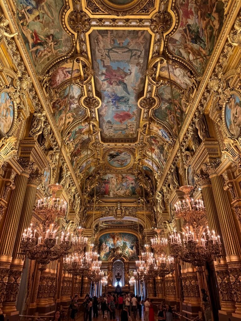 Salão ornamentado da Ópera Garnier com teto pintado de forma complexa, decorações douradas e grandes lustres; pessoas caminhando abaixo.