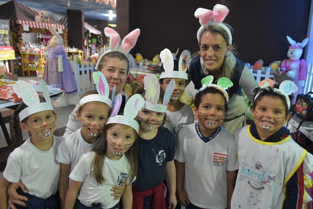 Na foto vemos 7 crianças com rostos pintados com nariz vermelho, bigode e dentinhos, usando orelhinhas de coelho feitas de papel, e duas mulheres adultas com orelhas de coelho de pelúcia. Foto facebook Vila Germânica.