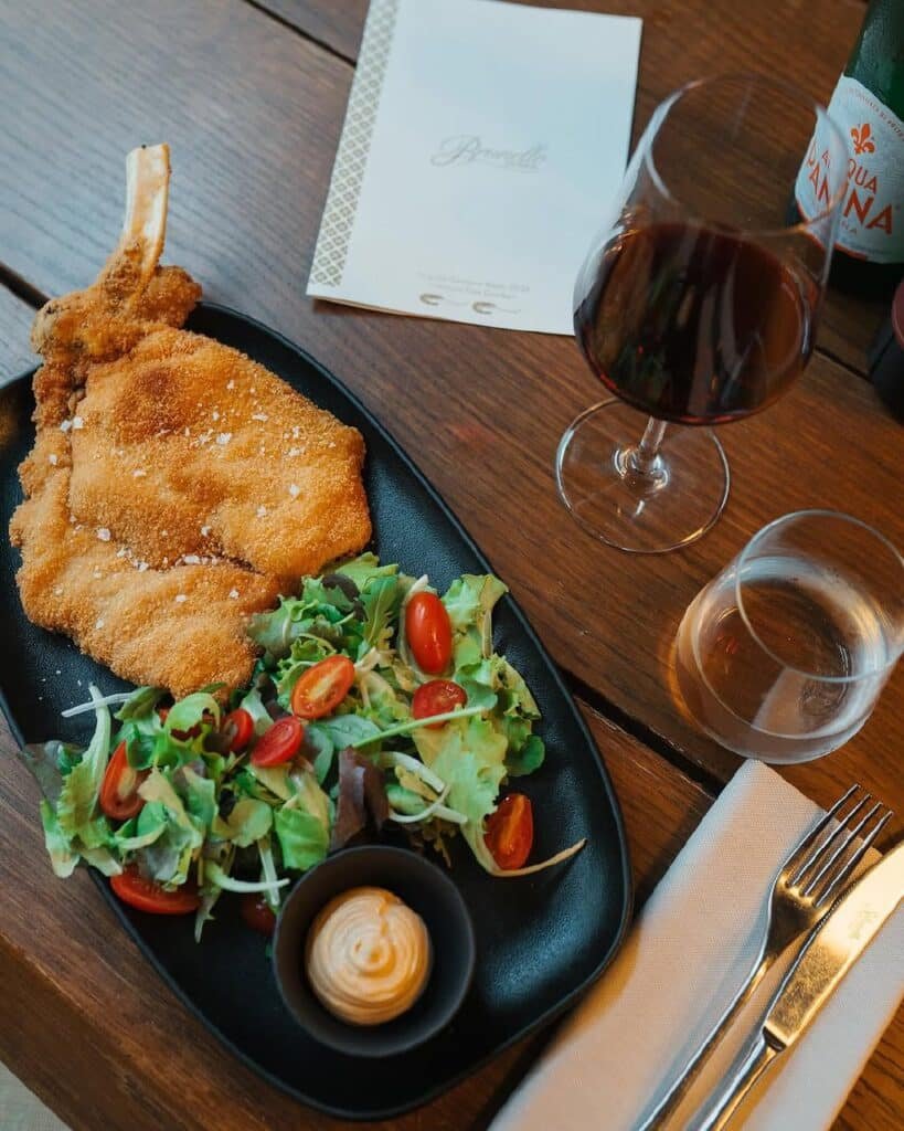Cotoletta alla Milanese (carne empanada, salada fresca com tomate cereja e molho para acompanhar) na Osteria Brunello. Uma taça de vinho tinto e um cardápio também estão na mesa.