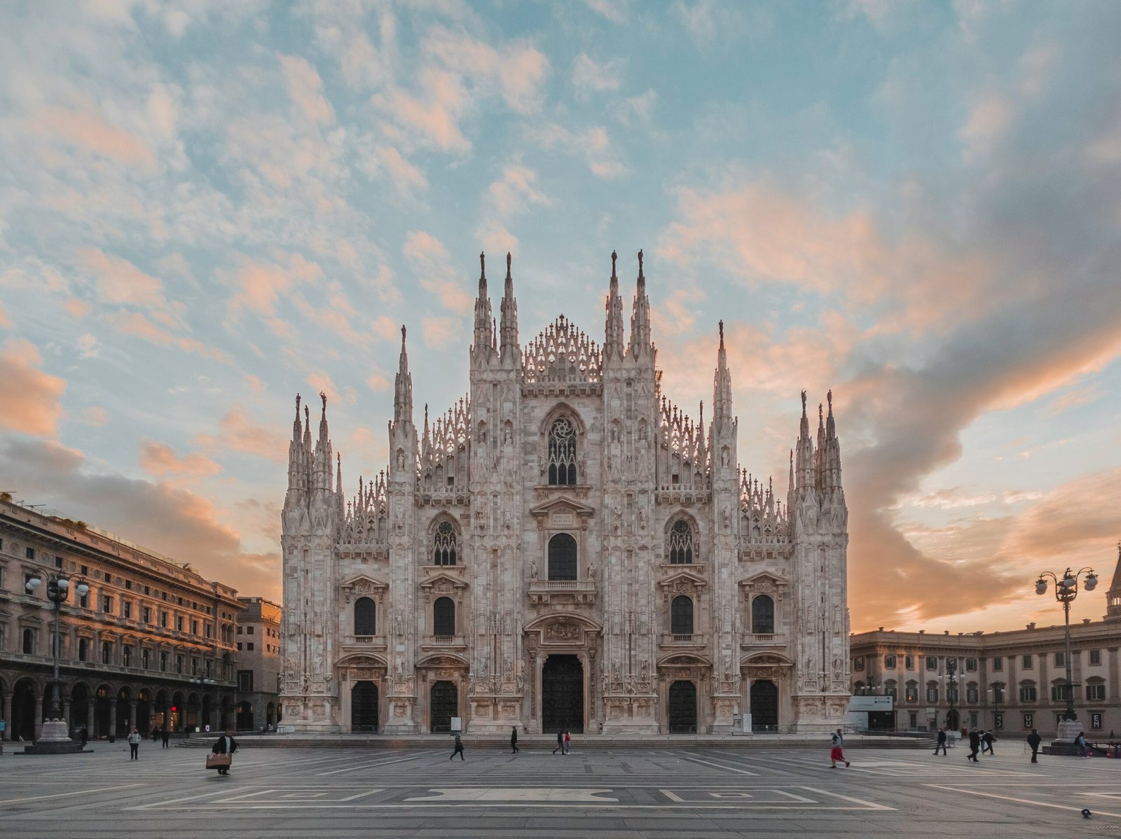 A imagem mostra a Catedral de Milão com sua fachada detalhada ao pôr do sol, em contraste com um céu parcialmente nublado e uma praça aberta. A foto está na lista de o que fazer em Milão.