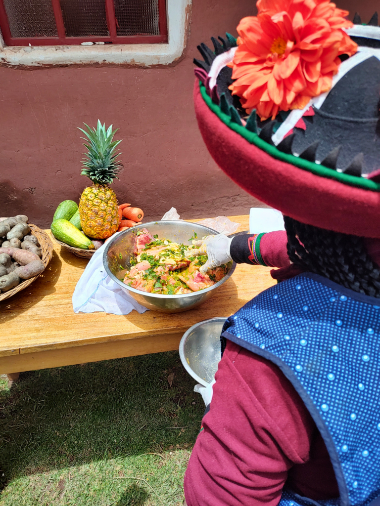 mesa de madeira com uma tigela de alumínio, onde há carnes cruas temperadas. No canto direito é possível ver metade de uma mulher de costas para a câmera, mexendo na tigela.