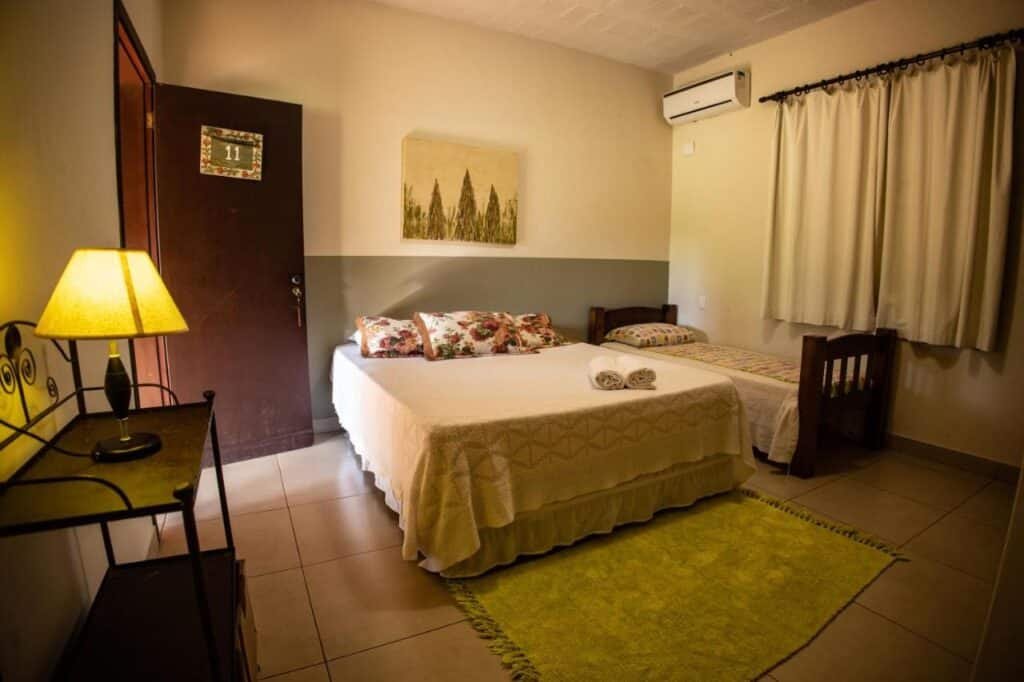 A imagem mostra um quarto simples e acolhedor do Paineiras Hotel Fazenda, com uma cama de casal e uma cama de solteiro. As camas são cobertas com colchas brancas, e sobre a cama de casal há almofadas decorativas florais. Uma luminária está acesa sobre uma pequena mesa de ferro ao lado da cama de casal. O quarto tem um tapete verde claro no chão e uma janela com cortinas bege ao lado da cama de solteiro. Acima da cama de casal, há um quadro com uma paisagem de árvores. O ambiente é iluminado e tem um ar-condicionado na parede.