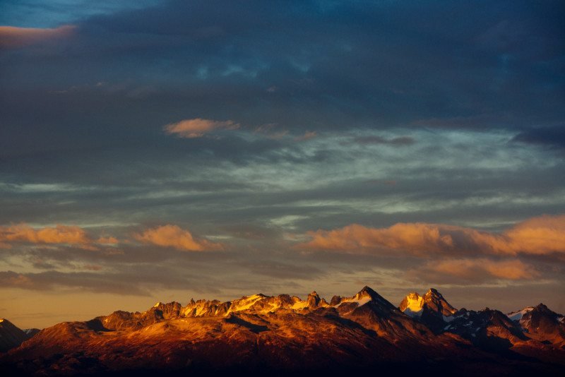 Uma cadeia de montanhas é banhada pela luz dourada do sol sob um céu com nuvens durante o pôr do sol. Imagem para ilustrar post sobre a Patagônia Argentina.