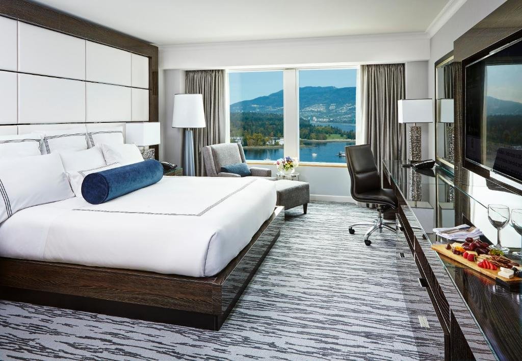 Quarto no Pan Pacific Vancouver com uma janela dando vista para a baía e montanhas, de frente para a janela há uma poltrona cinza com um abajur de chão ao lado. Tem uma cama de casal do lado esquerdo, com muitas almofadas sob a cama, na frente tem um rack com uma televisão