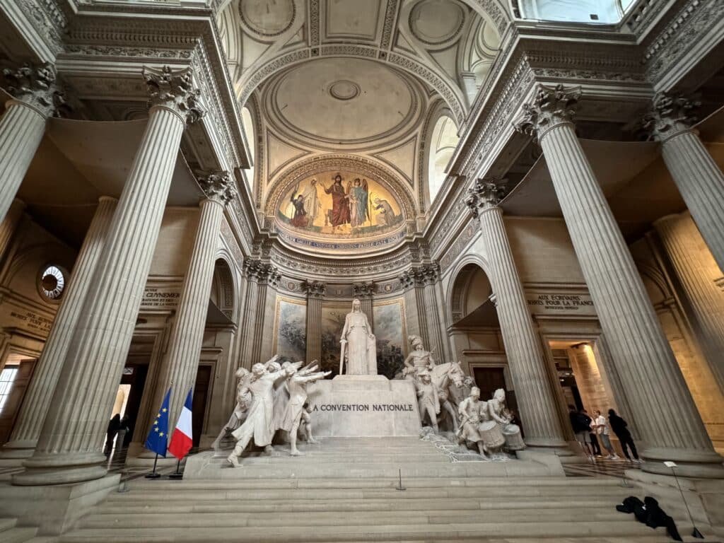 Interior de um grande salão com colunas altas, teto detalhado, uma estátua proeminente e bandeiras francesas. Visitantes ficam em pé ao redor da área interna do Panteão, um dos passeios em Paris.