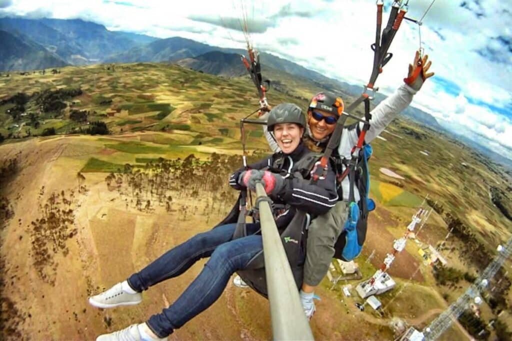 uma mulher toda equipada segundo um pau de selfie e fotografando ela mesma, junto ao instrutor de parapente atrás dela, voando sobre os ares, acima das planícies e montanhas do Vale Sagrado dos Incas. Ambos estão sorrindo para a câmera, a mulher é branca e o homem é moreno.