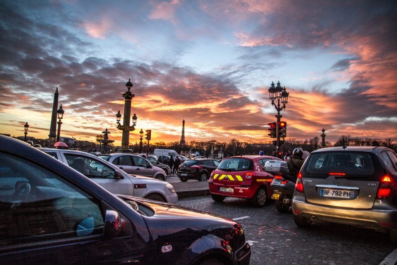 Engarrafamento ao pôr do sol com vários carros em uma rua de Paris. Postes de luz e um céu claro e nublado ao pôr do sol são visíveis ao fundo, assim como a Torre Eiffel.