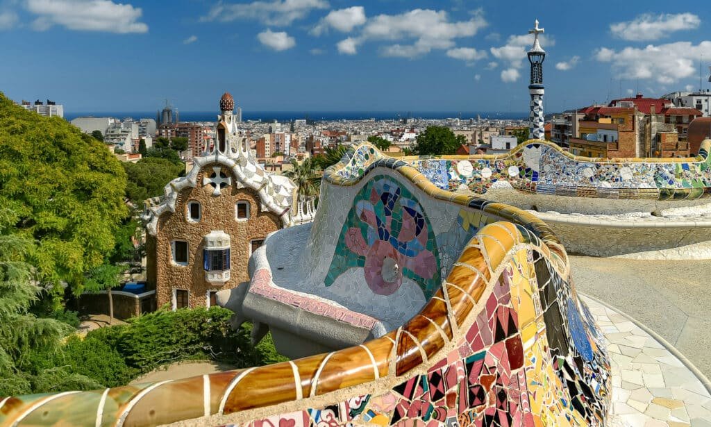 Vista do Parque Guell, em Barcelona, que pode ser visitado com o seguro viagem Espanha. Muros de azulejos coloridos ficam no centro da foto, e é possível ver casas e prédios da cidade ao fundo.